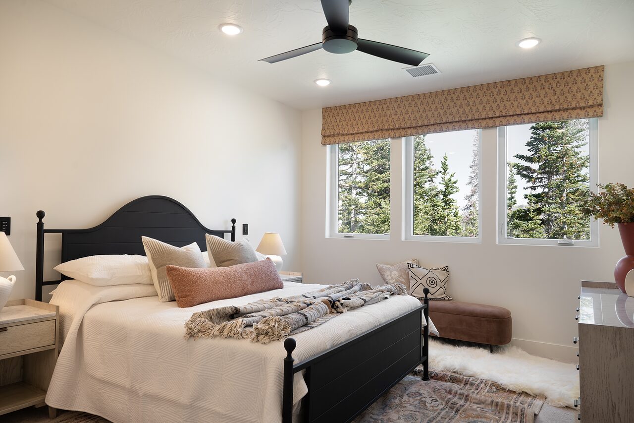 Primary bedroom with a black iron bed, layered cream and blush bedding, a woven roman shade, and forest-facing windows
