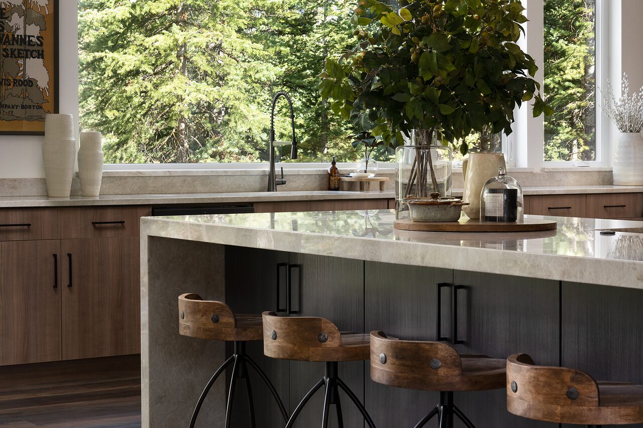 Kitchen island in veined pale stone with wood-seat black iron stools, warm wood cabinetry, a leafy branch in a vase, and pines through the window