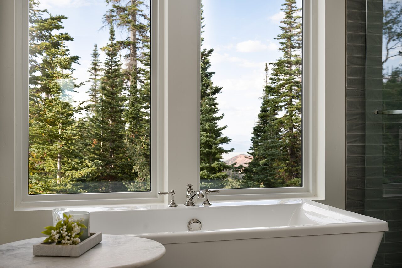 Freestanding white tub beneath tall windows framing evergreens, with a small potted plant on the ledge