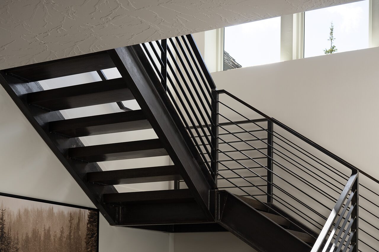 Black steel staircase with horizontal rails climbing past tall windows, forest light across the treads