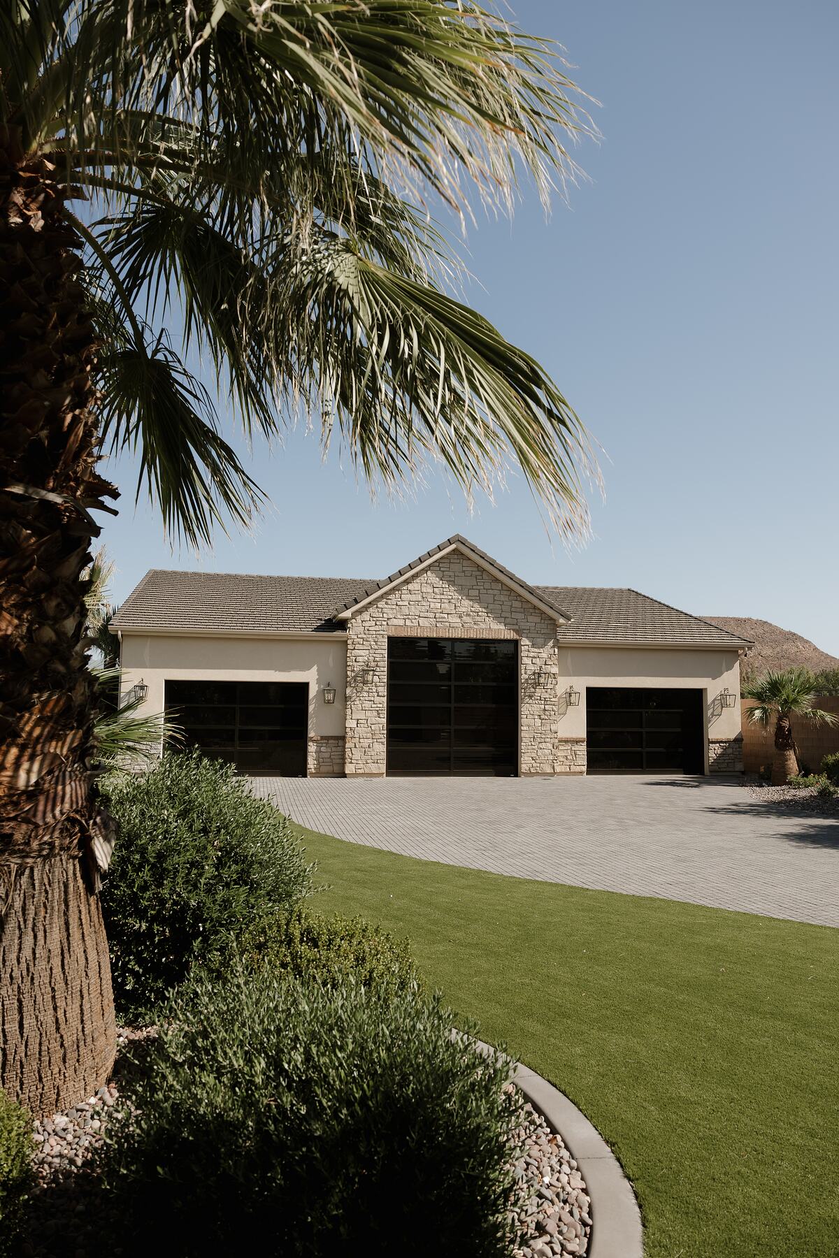 Three-bay garage with a stone-clad center gable, cream stucco wings, full-glass doors, lantern sconces, and a paver drive under a palm