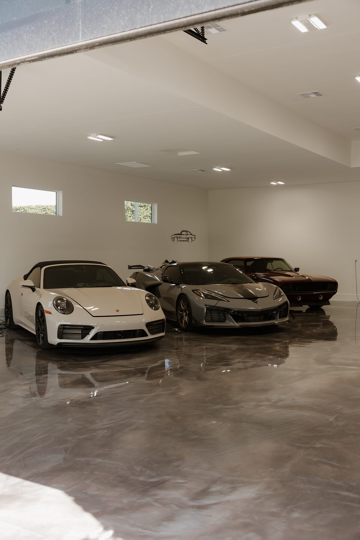 Garage interior with a polished poured floor reflecting a white sports car, a gray coupe, and a dark classic, under a bright recessed-lit ceiling