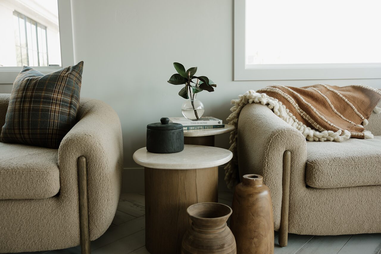 Two camel boucle armchairs flanking a travertine side table with a black lidded vessel, wooden vases, and a tan fringed throw