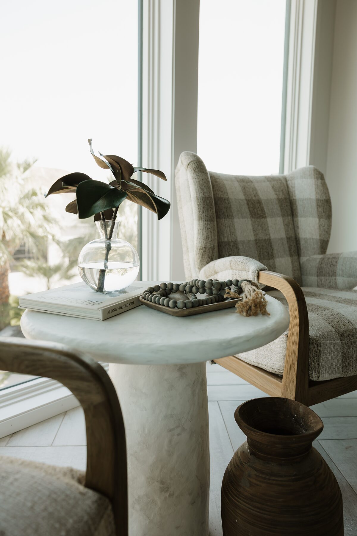 Gingham wingback chair beside a plaster pedestal table holding a magnolia stem in a glass vase, wood beads, and a book