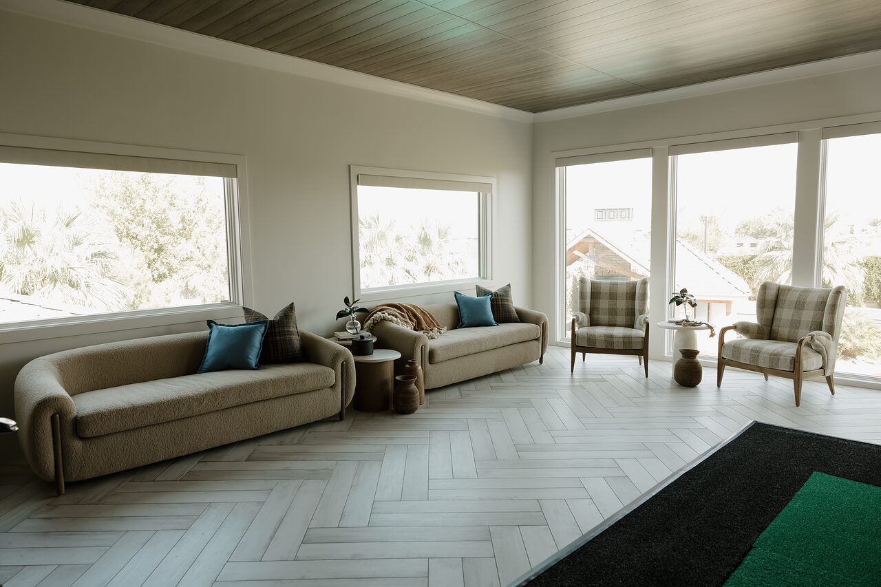 Wide view of the lounge with two sofas, blue pillows, gingham chairs at the windows, and the green hitting mat in the foreground