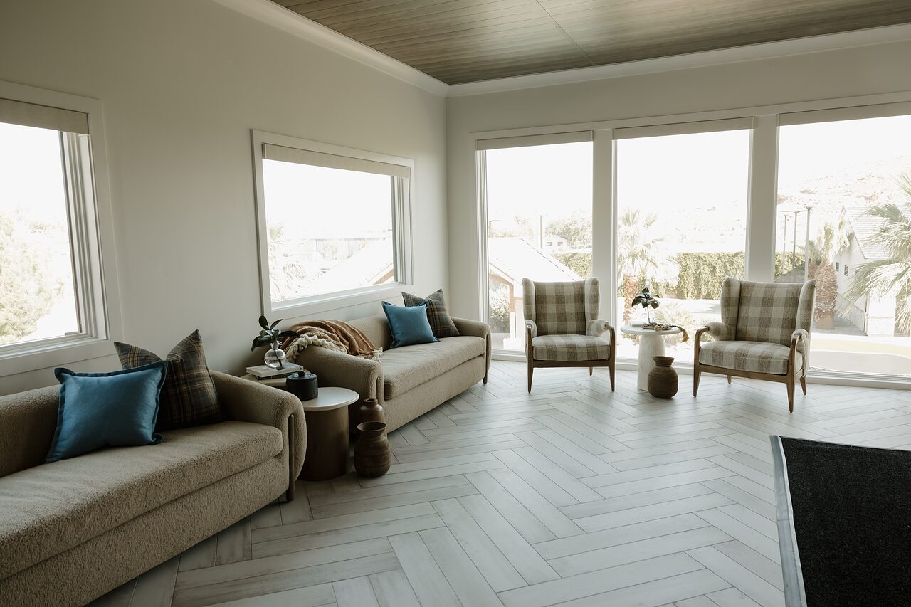 Sunlit lounge with two camel boucle sofas, gingham wingback chairs, a herringbone tile floor, and windows on three sides