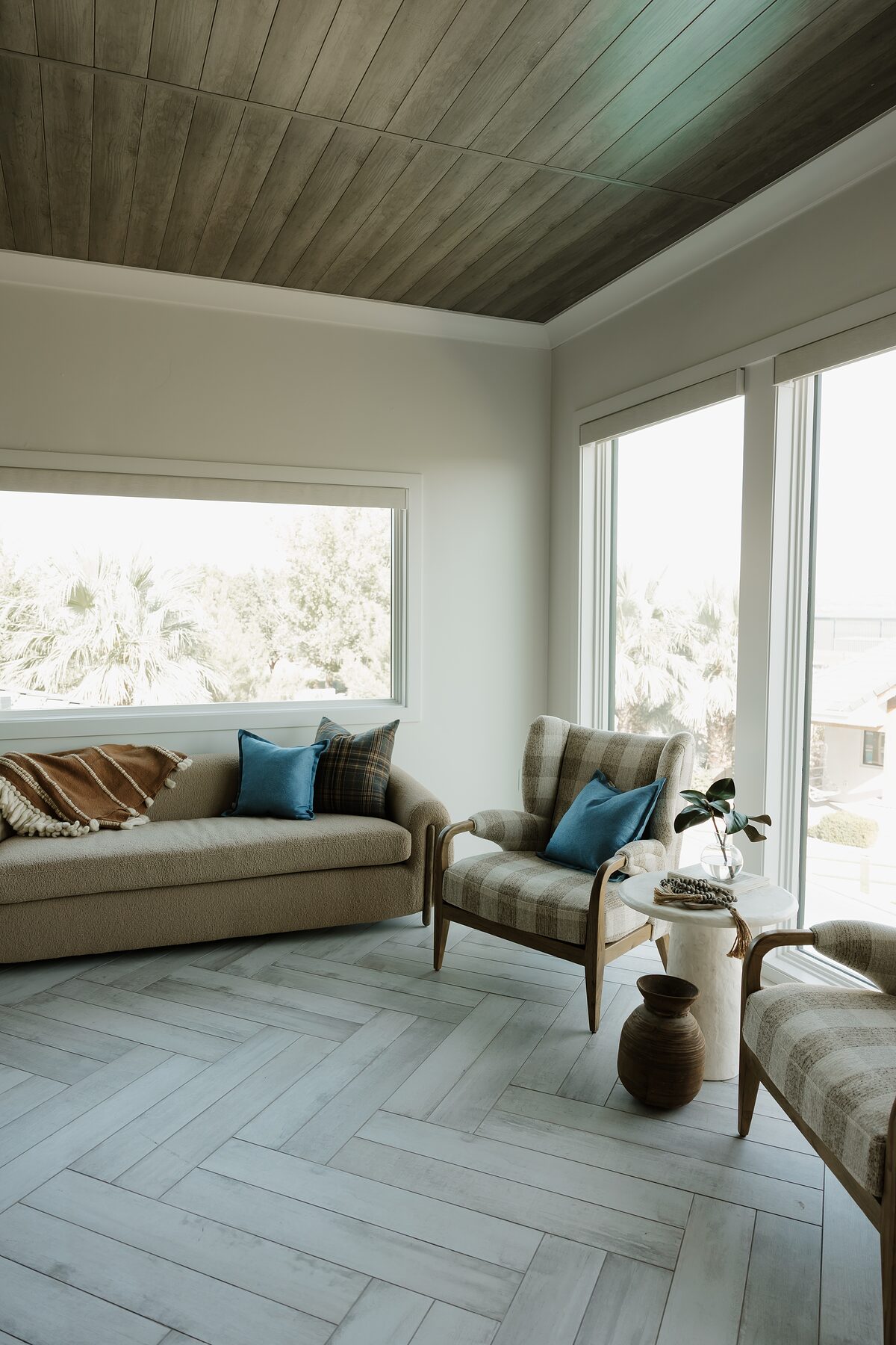 Corner of the lounge showing the gray plank ceiling, a boucle sofa with blue and plaid pillows, and a gingham wingback by the window