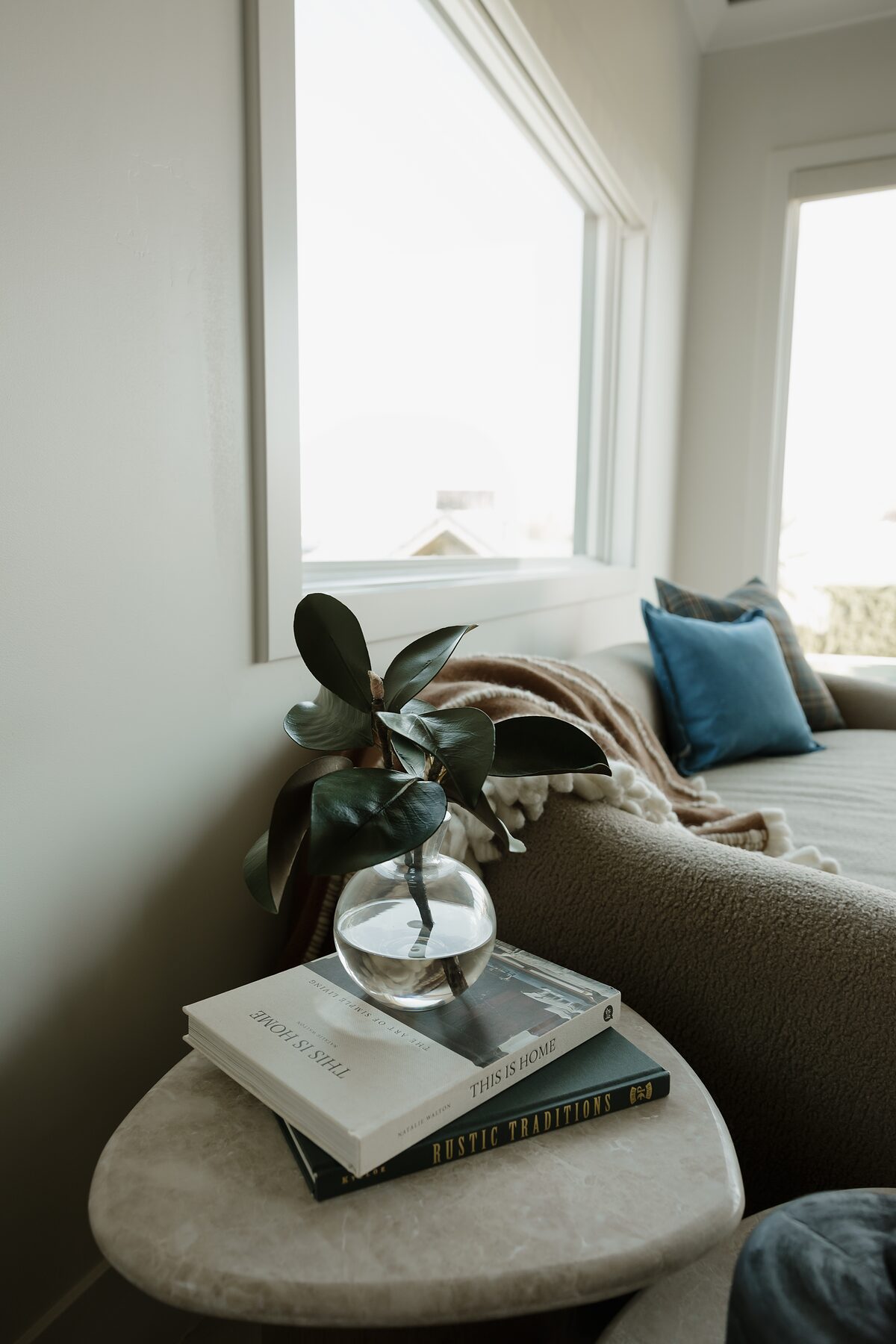 Stack of design books and a glass bud vase with rubber plant leaves on a stone side table beside the sofa