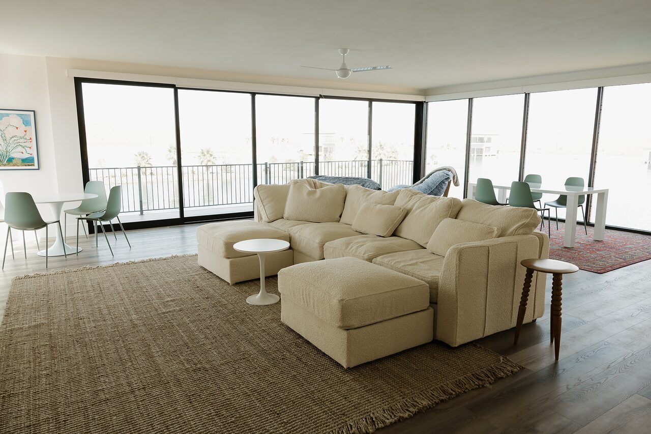Boathouse great room with a cream sectional on a jute rug, sage dining chairs, and floor-to-ceiling glass looking onto the water
