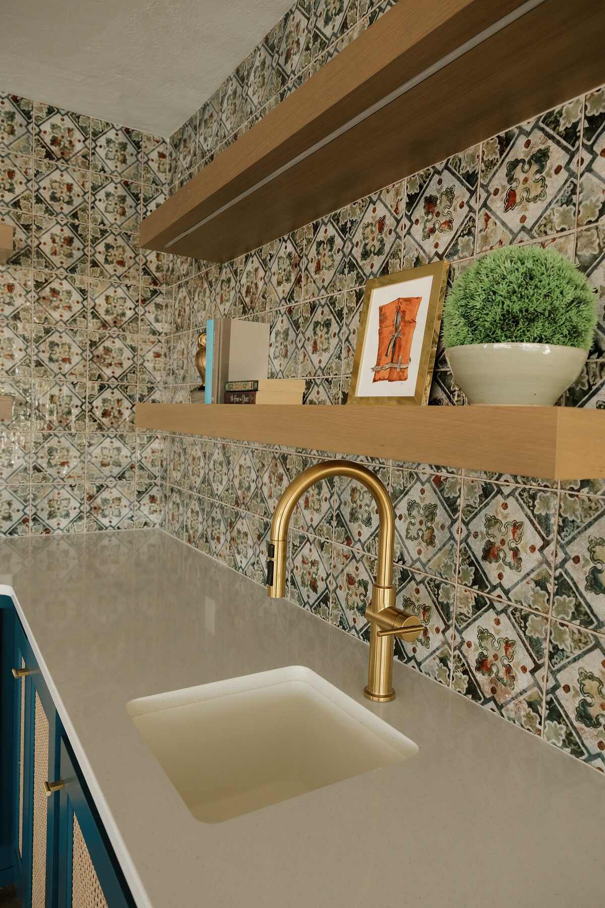 Wet bar detail with patterned green and rust tile, floating oak shelves, a brushed brass faucet, and teal cabinets